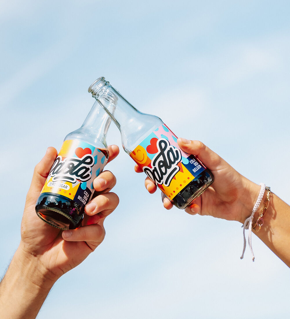 Deux personnes trinquant avec des bouteilles de soda local Paola, sous un ciel bleu